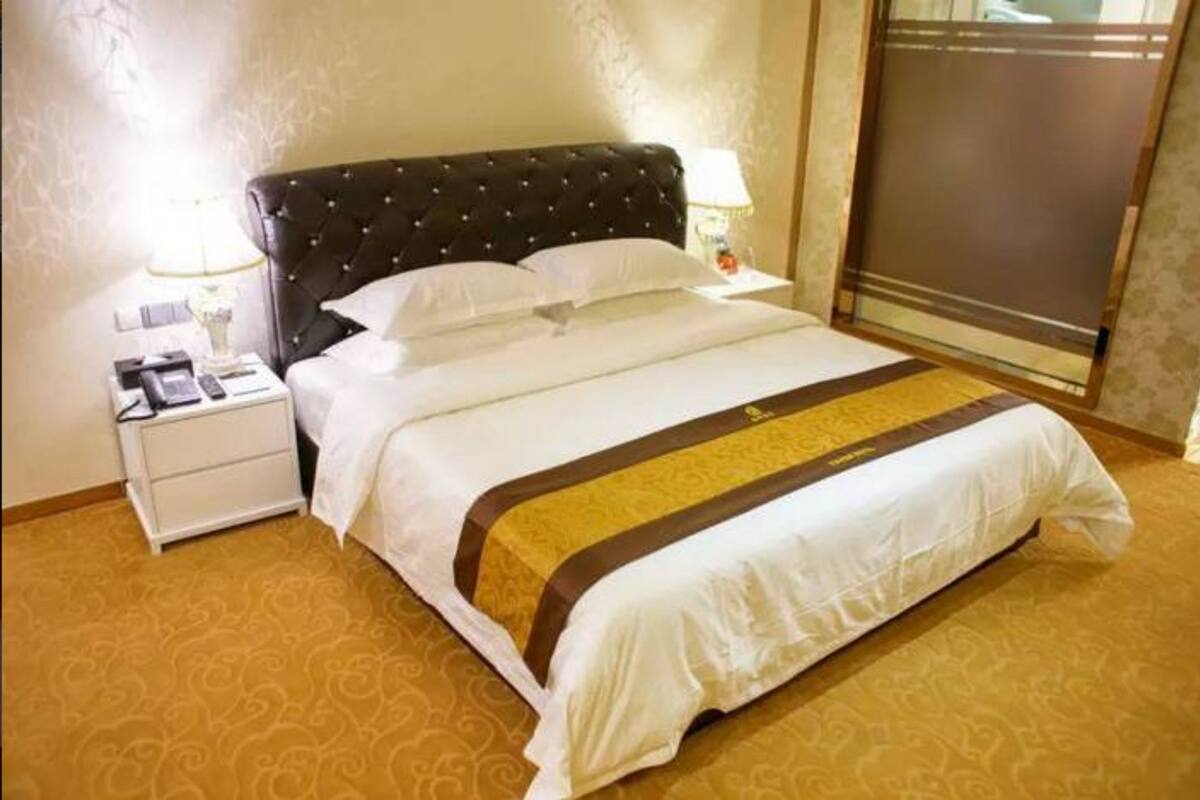 Cama bem arrumada com cabeceira estofada, roupa de cama branca e um corredor de cama decorativo dourado e castanho. Há uma mesa de cabeceira com uma luminária e um telefone ao lado da cama.