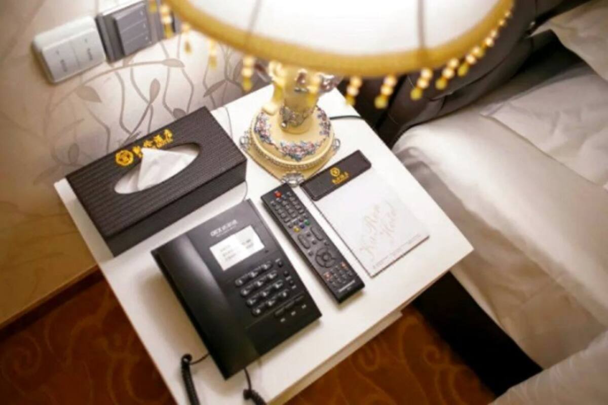 Quarto de hotel bem arrumado com uma mesa de cabeceira que contém uma lâmpada, um controle remoto, uma caixa de lenços, uma carteira preta e um pequeno livreto.