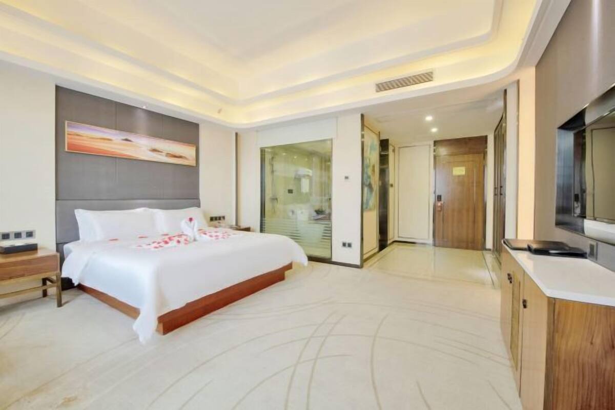 A modern hotel room with a large bed, a television, a desk, and a bathroom visible through a glass door.