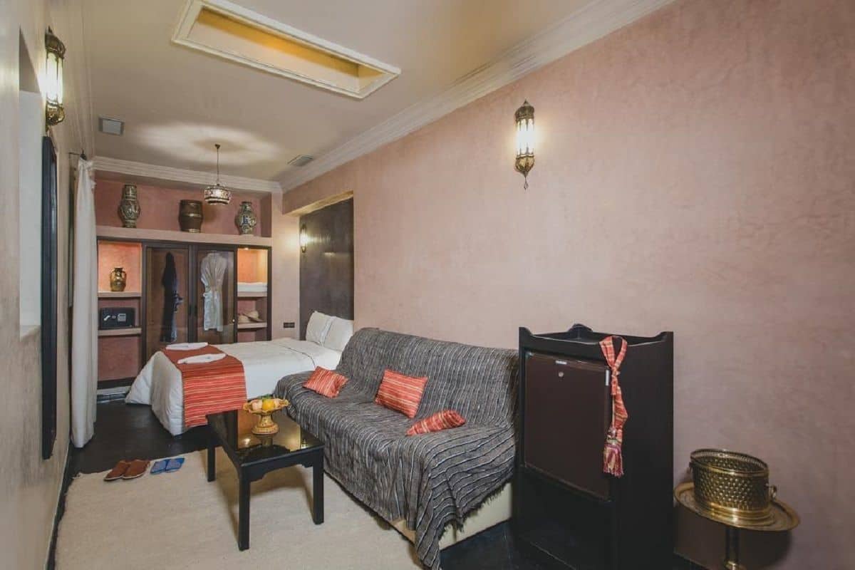 A hotel room with a bed, a sofa, a small table, and a cabinet. There is a window with curtains, a wall-mounted lamp, and a ceiling light.