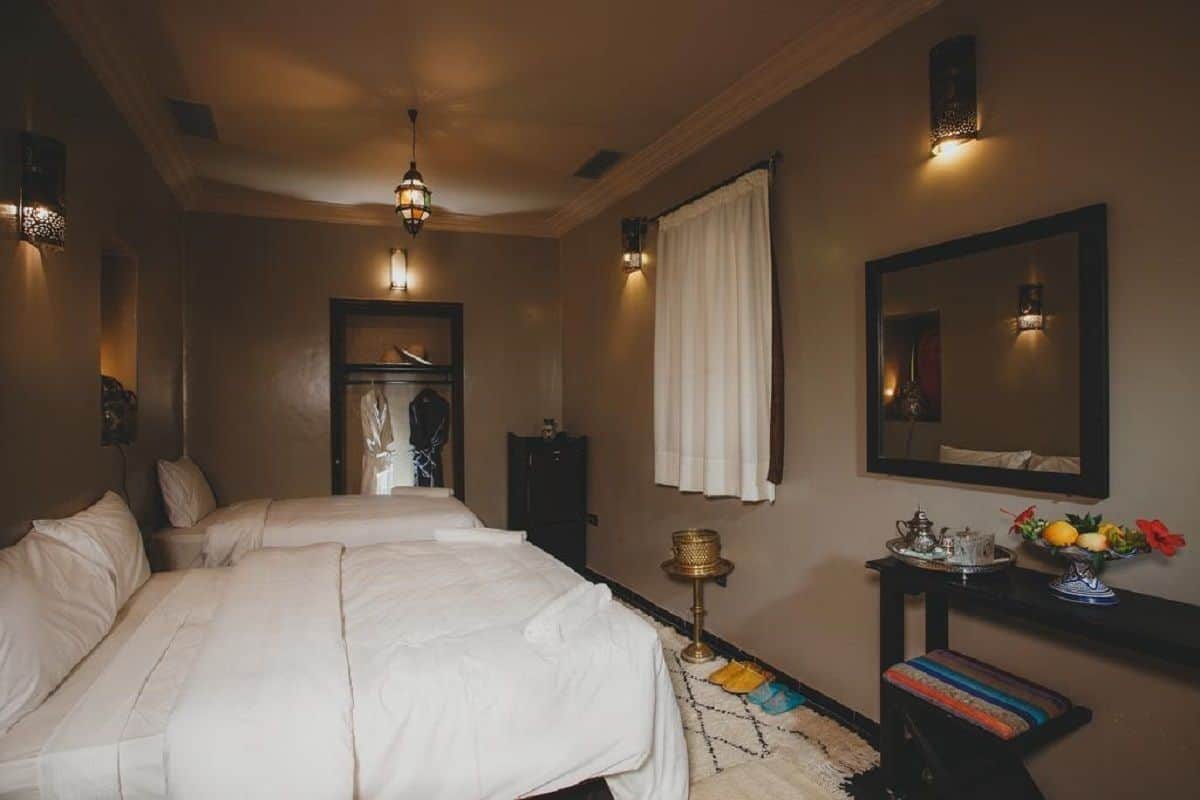 A hotel room with two beds, a wardrobe, a mirror, a nightstand with a lamp, and a small table with a vase of flowers.
