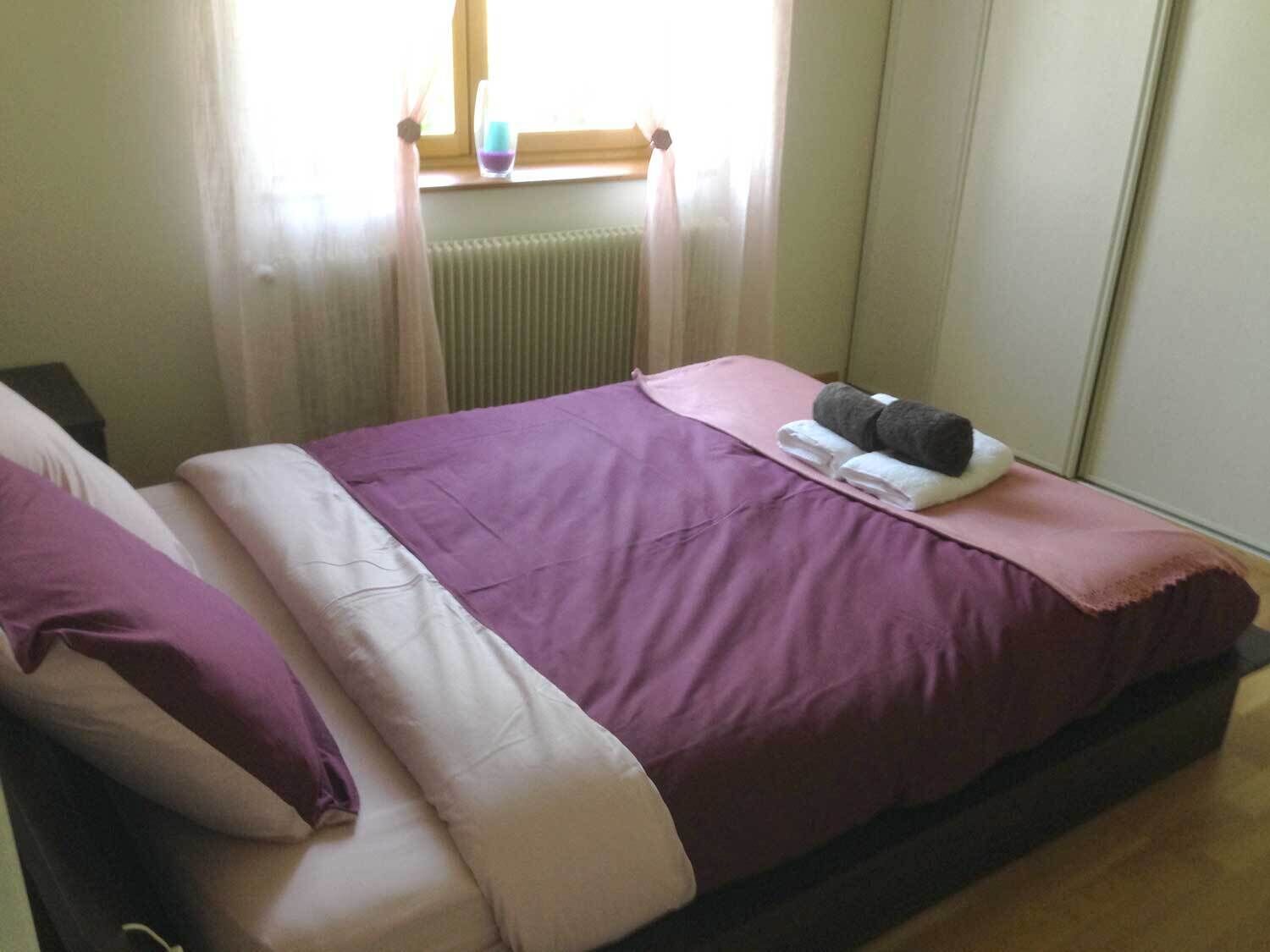 Een net opgemaakt bed met een paars dekbed en witte kussens, een raam met dunne gordijnen en een radiator onder het raam.