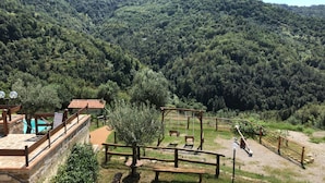 A rustic wooden deck with a stone wall, a wooden swing set, and a pool area with a tiled roof. - Agriturismo Piano di Andrea (Perito)