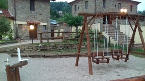A rustic outdoor area with a wooden swing set, stone walls, and a building with a tiled roof in the background. - Agriturismo Piano di Andrea (Perito)