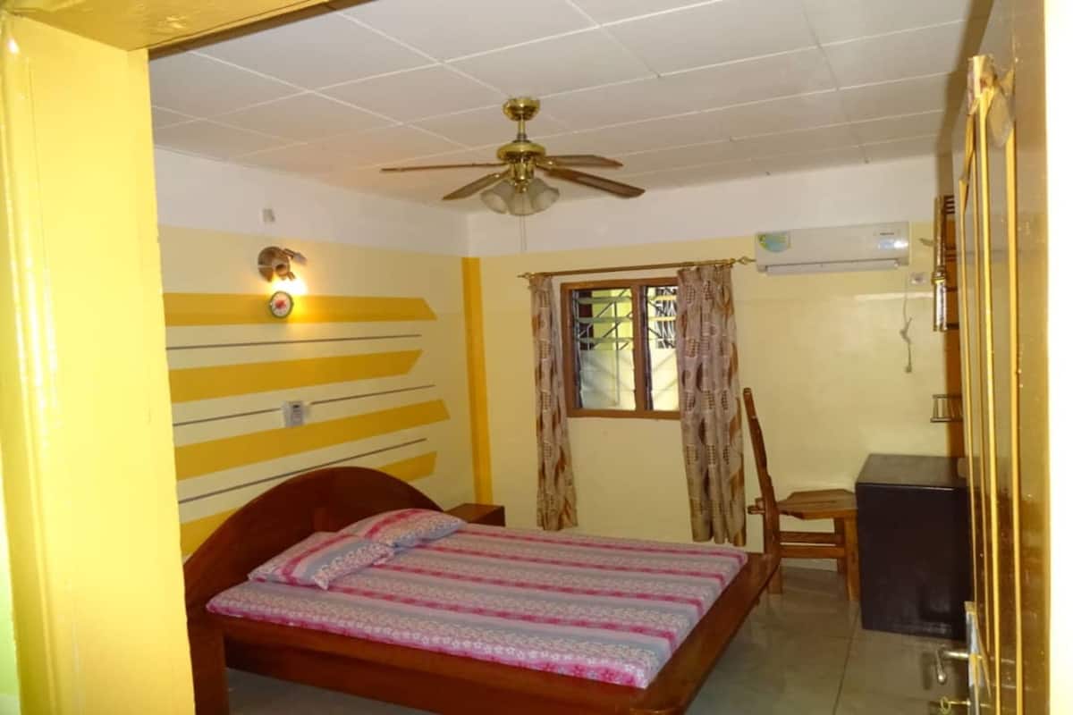 Une chambre à coucher comprenant un lit, un bureau, une chaise, un ventilateur de plafond et une fenêtre avec des rideaux.