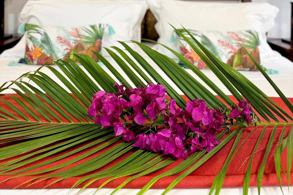 A bed with a tropical-themed pillow, a painting of a bridge in a forest, and a bouquet of purple flowers.