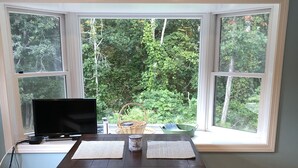 A dining area with a wooden table and bench, a TV, and a view of greenery through large windows.