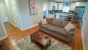 A living room with a sofa, coffee table, and a rug. There is a kitchen in the background with cabinets and appliances.