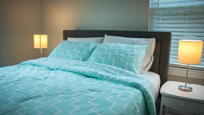 A neatly made bed with a patterned bedspread and pillows, a bedside table with a lamp, and a window with blinds.