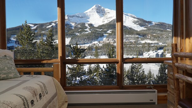 A cozy cabin room with a large window offering a view of a snow-covered mountain and forest. - SPECTACULAR VIEW of Big Sky-Hot Tub & Fireplace-1/2 Mile on Free Shuttle to Mtn. (Big Sky)