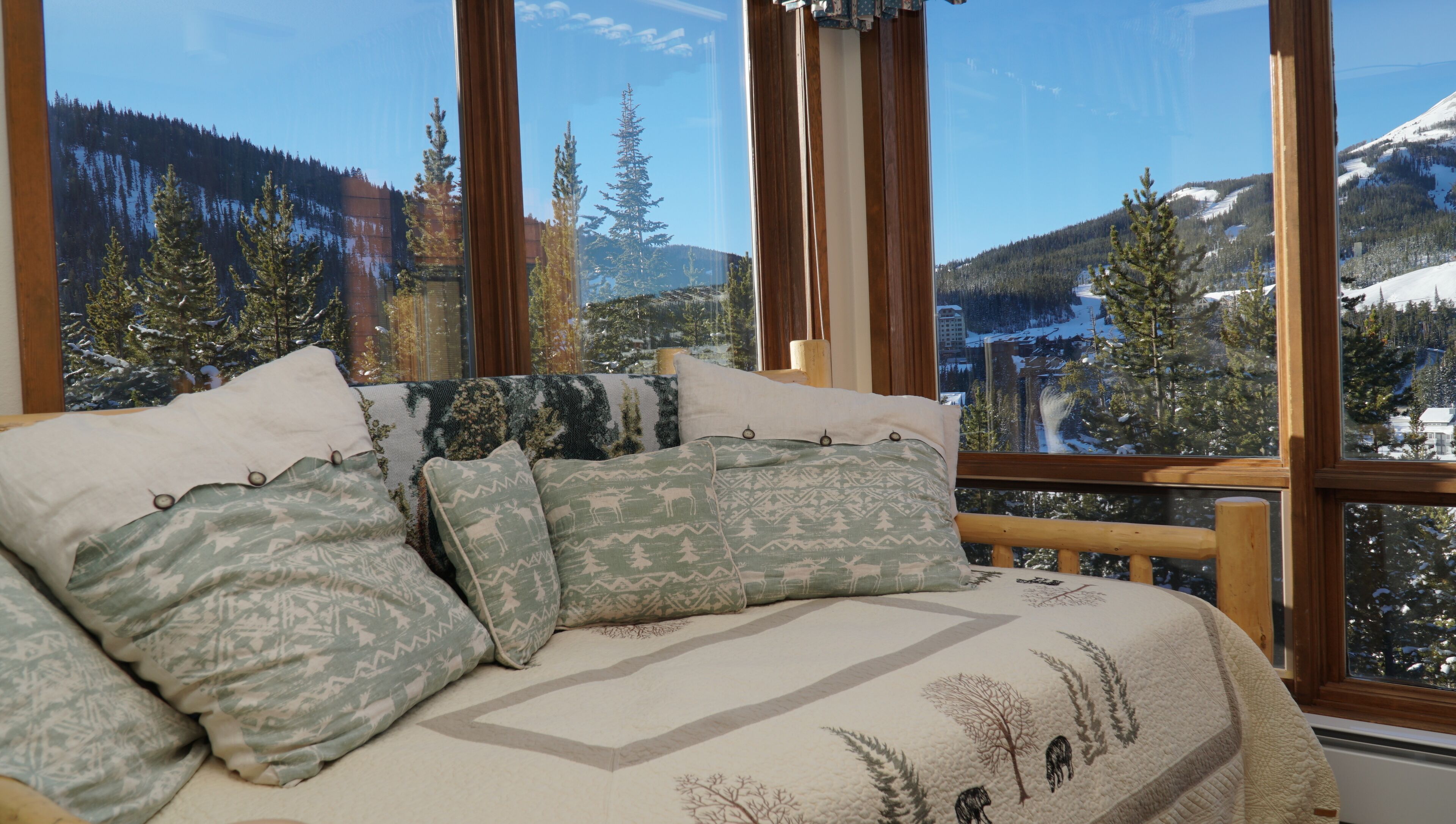 Ein Bett mit gemusterten Bettwäsche und Kissen, ein Holz-Kopfteil und ein Blick auf eine verschneite Berglandschaft durch große Fenster.
