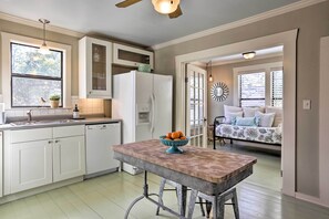 Una cocina con armarios blancos, un refrigerador blanco, una mesa de madera con un cuenco de fruta y una ventana con vistas al exterior.