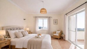Une chambre à coucher comprenant un lit, des tables de chevet, un luminaire suspendu, une fenêtre avec des rideaux et une vue sur l’extérieur.