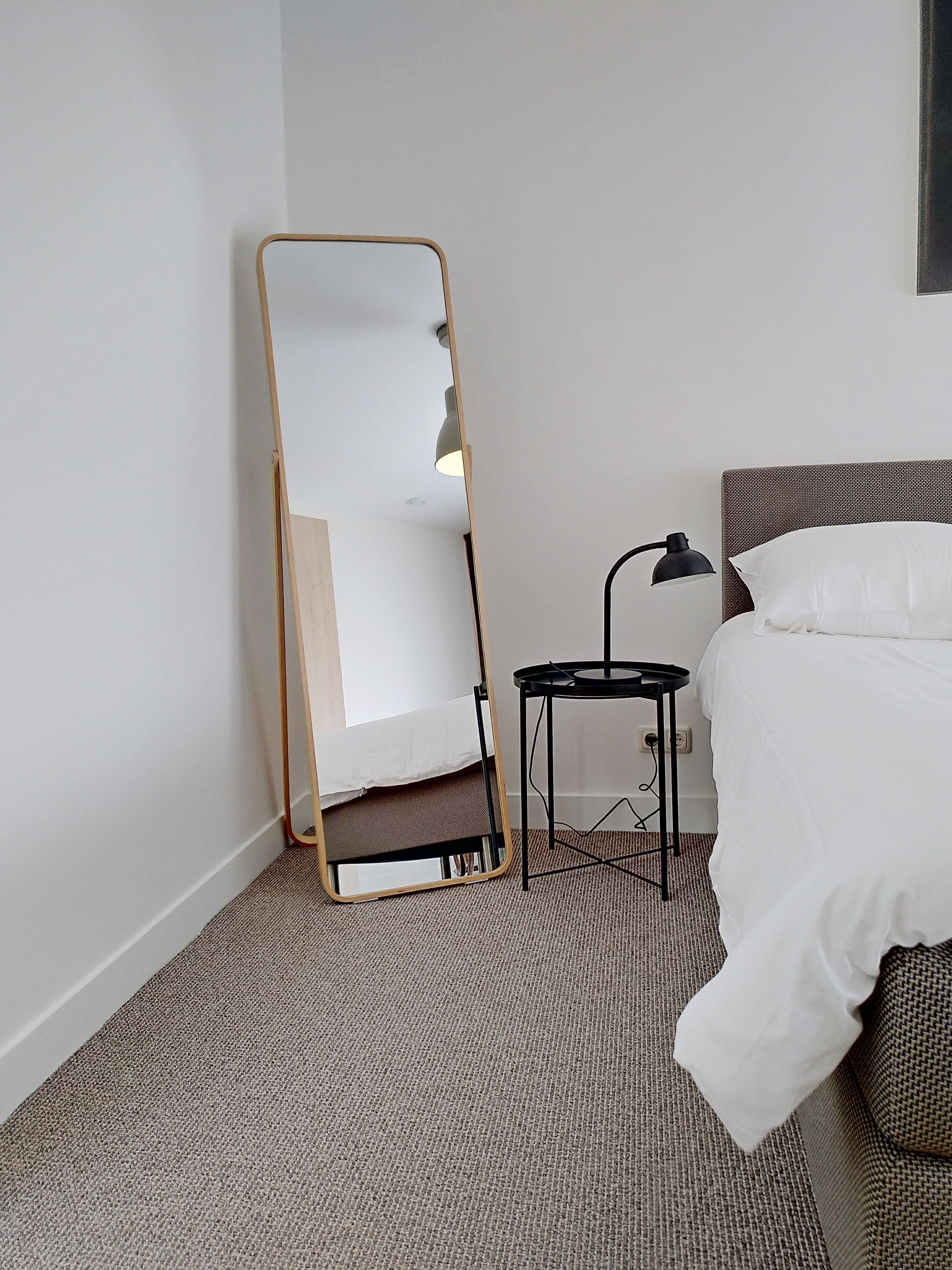 A bedroom with a bed, a bedside table with a lamp, and a full-length mirror leaning against the wall.