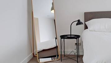 A bedroom with a bed, a bedside table with a lamp, and a full-length mirror leaning against the wall.
