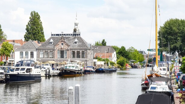 Un canal où sont amarrés des bateaux, un bâtiment historique portant une enseigne « DE VOLHARDING », et un grand mât.
