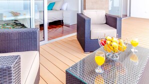 A modern outdoor seating area with wicker furniture, a glass table with fruit and drinks, and a view into a room with a bed and a painting. - Villa Chimera by LovelyStay (Caniço)