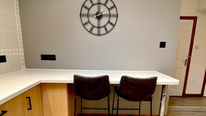 A kitchen with a white countertop, two bar stools, a wall-mounted clock, and wooden cabinets. - 3 Sheraton Apartments, Modern Luxury Apartment,Breathtaking Views of Ben Nevis (Fort William)