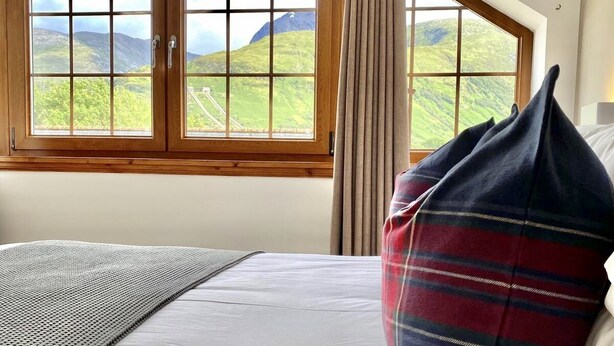 A bedroom with a large window, a bed with white bedding, a plaid pillow, and a textured throw blanket. - 3 Sheraton Apartments, Modern Luxury Apartment,Breathtaking Views of Ben Nevis (Fort William)