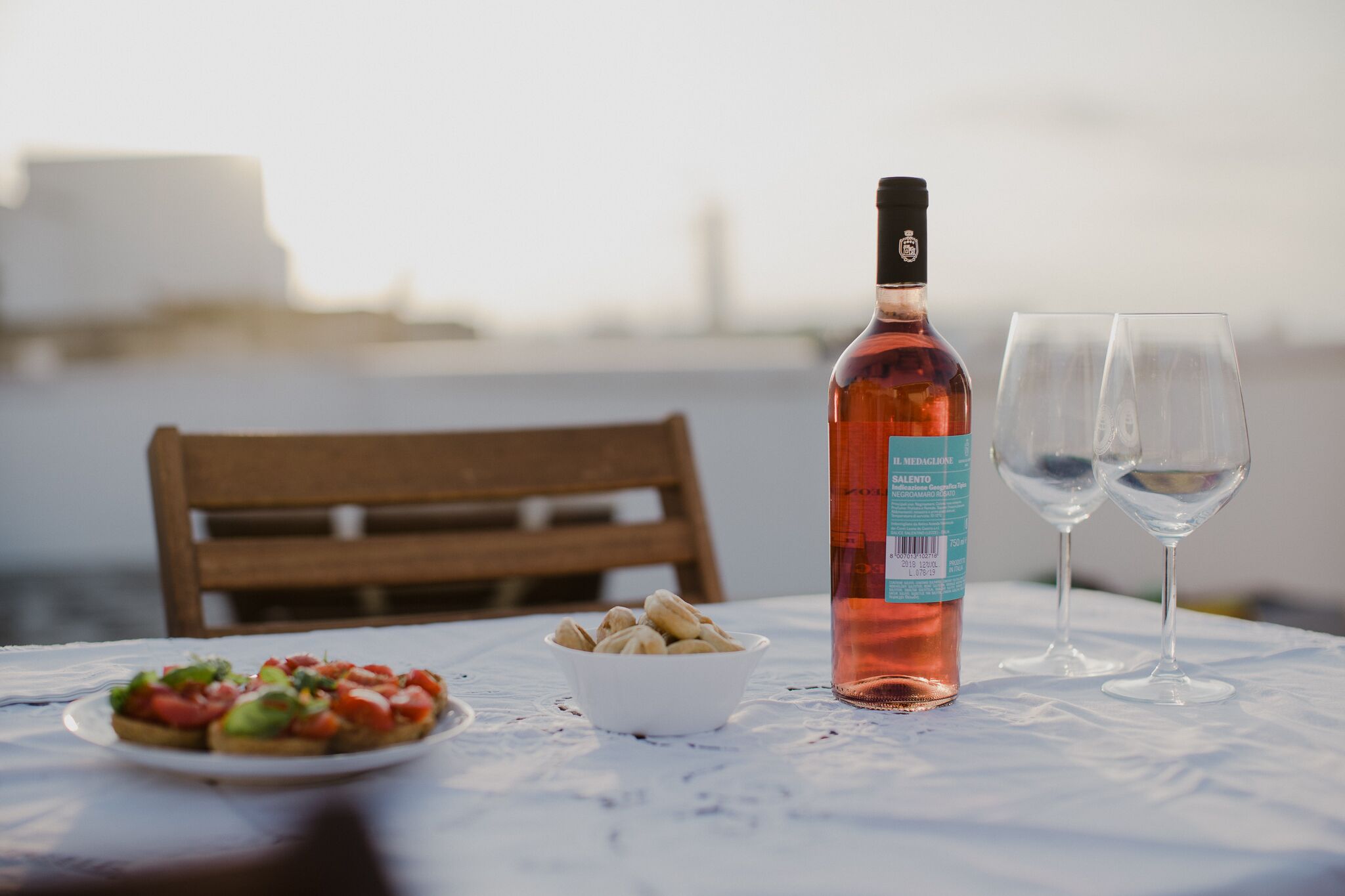 Una mesa con una botella de vino rosado, dos copas de vino, un bol de frutos secos y una fuente de bruschetta.
