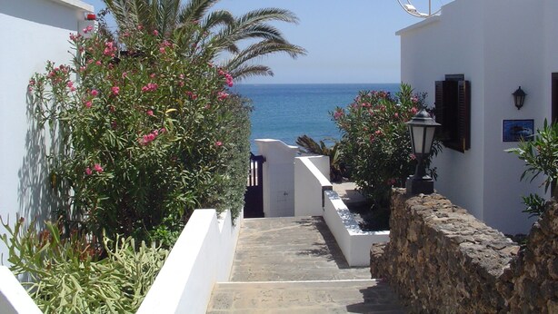 A narrow, white-walled pathway leading to a stone wall with a view of the sea.