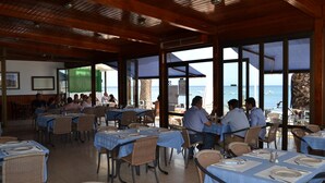 A restaurant with tables set for dining, large windows offering views of the sea, and a wooden ceiling.