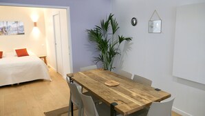 A room with a wooden dining table, white chairs, a bed with white and orange pillows, a large green plant, and a wall-mounted clock. - CENTRAL = BEAUTIFUL Area ; MODERN BRIGHT Apartment (Paris)