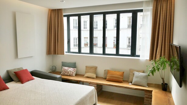 A bedroom with a bed, a bench with pillows, a window with curtains, and a potted plant. - CENTRAL = BEAUTIFUL Area ; MODERN BRIGHT Apartment (Paris)
