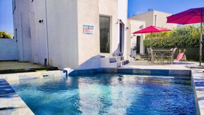 Una piscina con agua azul cristalina, rodeada por un edificio blanco con escalones que conducen a una puerta. Se observan sombrillas rojas y un tobogán al fondo.