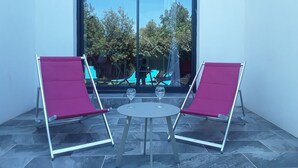 Un patio con dos sillas rojas y una mesita, una puerta corrediza de vidrio y vistas a árboles y una piscina azul.