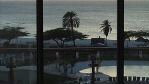 A view of the ocean from a window, with palm trees and a pool area visible. - Luxury, Eagle Beach Front 2BR, Penthouse  Private Roof Terrace (Eagle Beach)