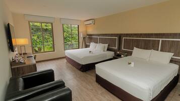 Habitación de hotel con dos camas, un escritorio con lámpara, un televisor y ventana con vista a follaje.