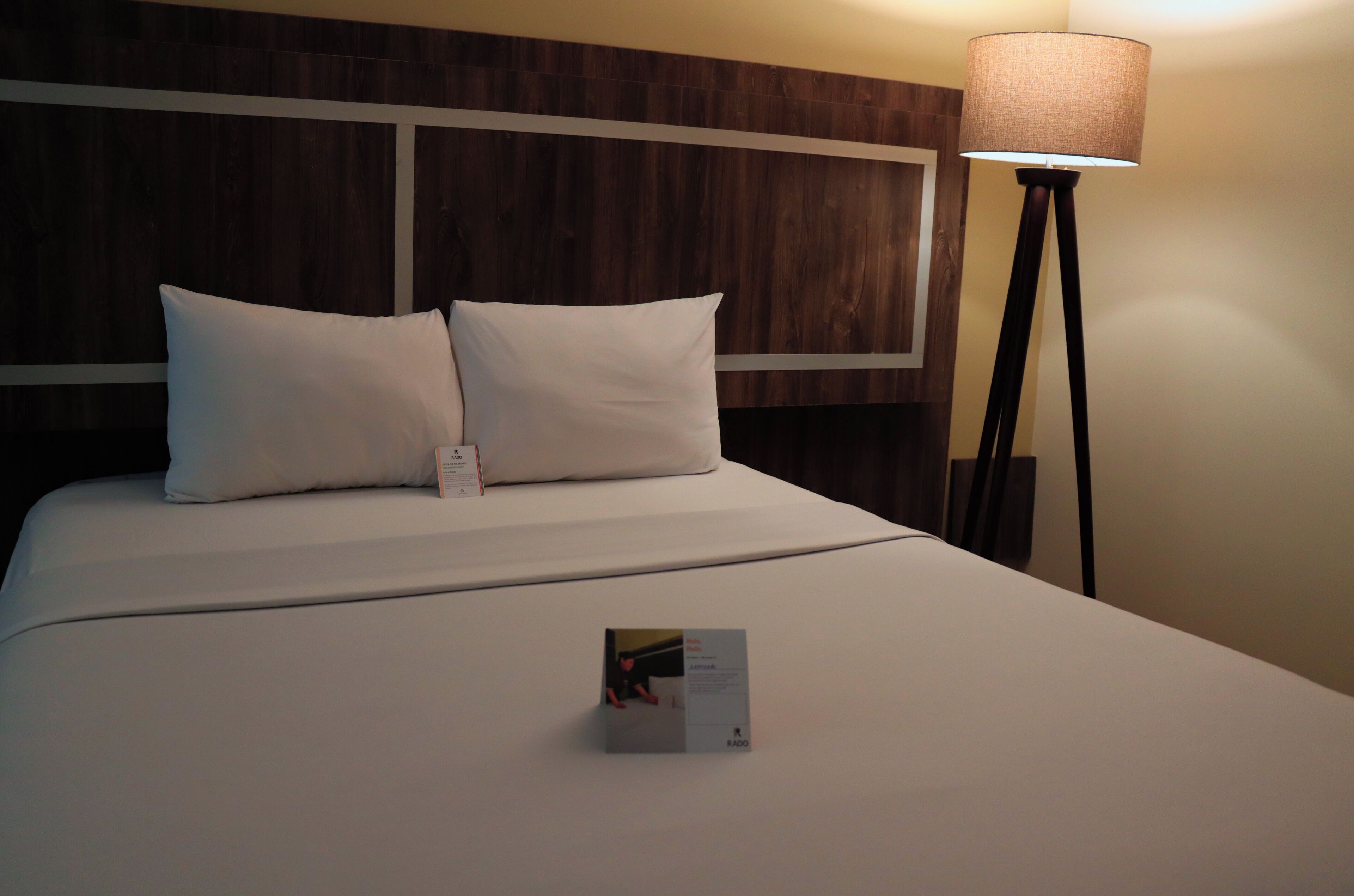 Een keurig opgemaakt bed met witte lakens en kussens, een nachtkastje met een lamp, en een klein informatief kaartje op het bed.