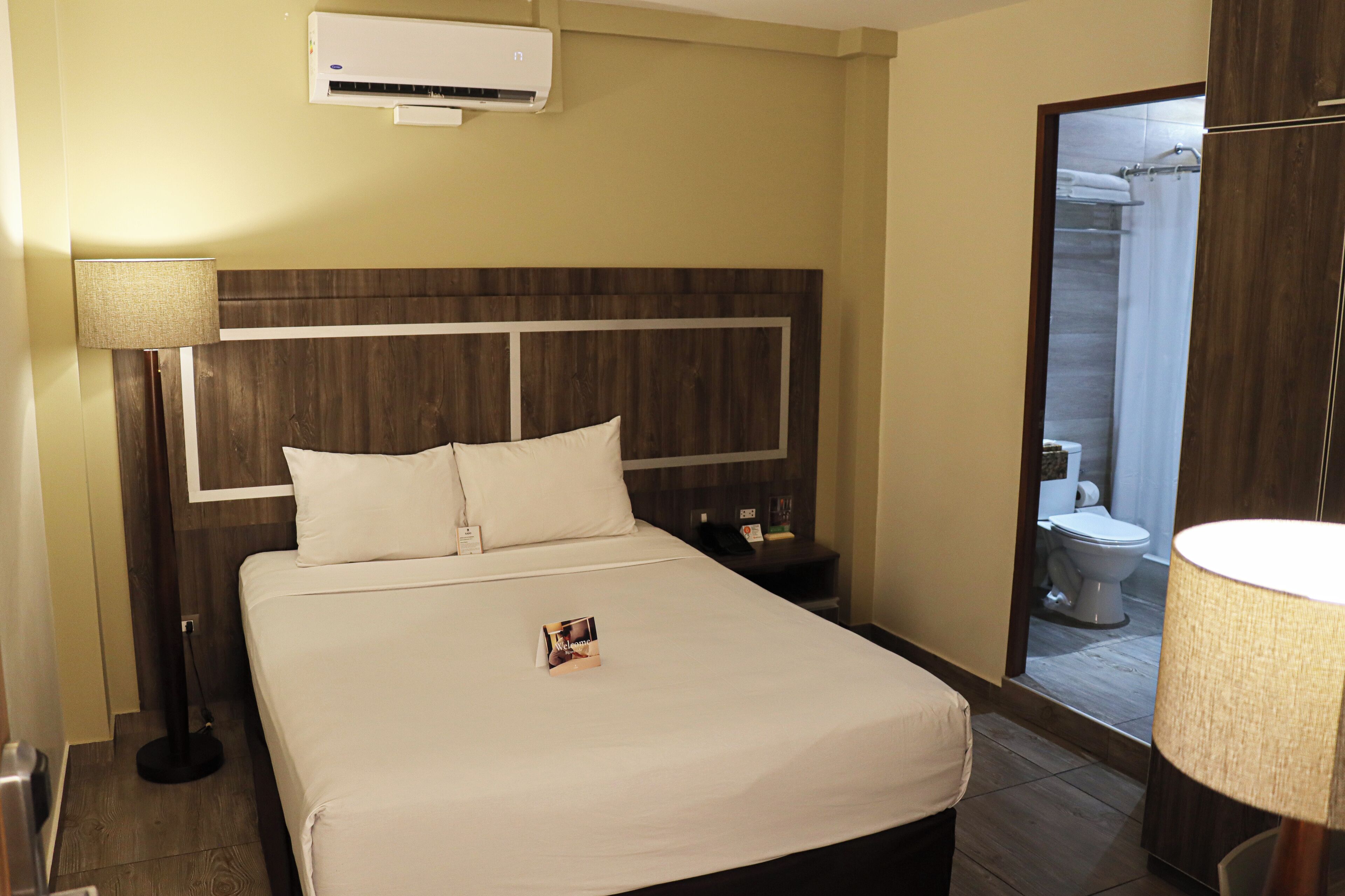 A hotel room with a bed, a nightstand, a lamp, an air conditioner, and a view of the bathroom.