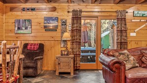 A wooden cabin interior with a leather couch, a chair with a red blanket, a small table with a lamp, and a door with curtains.