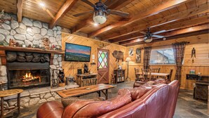 A cozy lodge interior with a stone fireplace, leather sofa, wooden ceiling beams, and a dining area.
