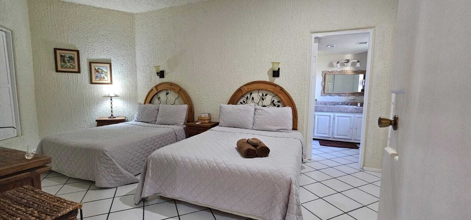 A hotel room with two beds, a wooden headboard, a small bedside table, a lamp, and a bathroom visible through an open door.