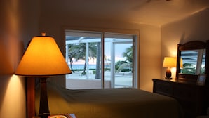 A hotel room with a bed, a lamp, a dresser, and a view of the beach through the window. - Sea Paradise # 24 (Cayman Islands)