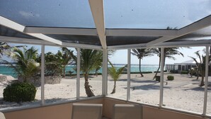 A covered outdoor seating area with a view of a sandy beach and the ocean. - Sea Paradise # 24 (Cayman Islands)
