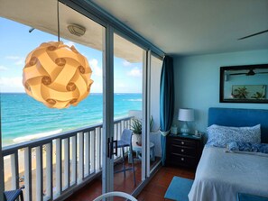 Een balkon met uitzicht op de oceaan, een bed met een blauw hoofdeinde, een nachtkastje, een lamp en een stoel.