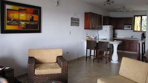 Una sala de estar con un sofá, una silla y una mesa de comedor con sillas. Hay una zona de cocina con armarios y un microondas. Se ve un ventilador de techo, y un cuadro cuelga en la pared.