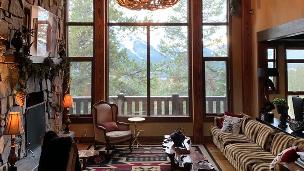 A cozy living room with a stone wall, a chandelier, a fireplace, and a large window with a view of mountains. - One Of A Kind Mountain Home With Gorgeous  mountain Views  (Canmore)