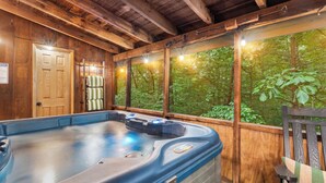 Une cabane en bois dotée d’un bain à remous, de murs en bois et offrant une vue sur une forêt grâce à de grandes fenêtres.