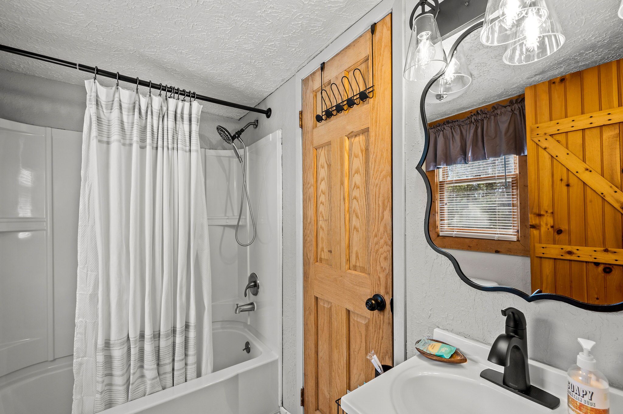 Une salle de bain avec un rideau de douche, une porte en bois, un lavabo avec un distributeur de savon et un miroir avec un cadre en bois.