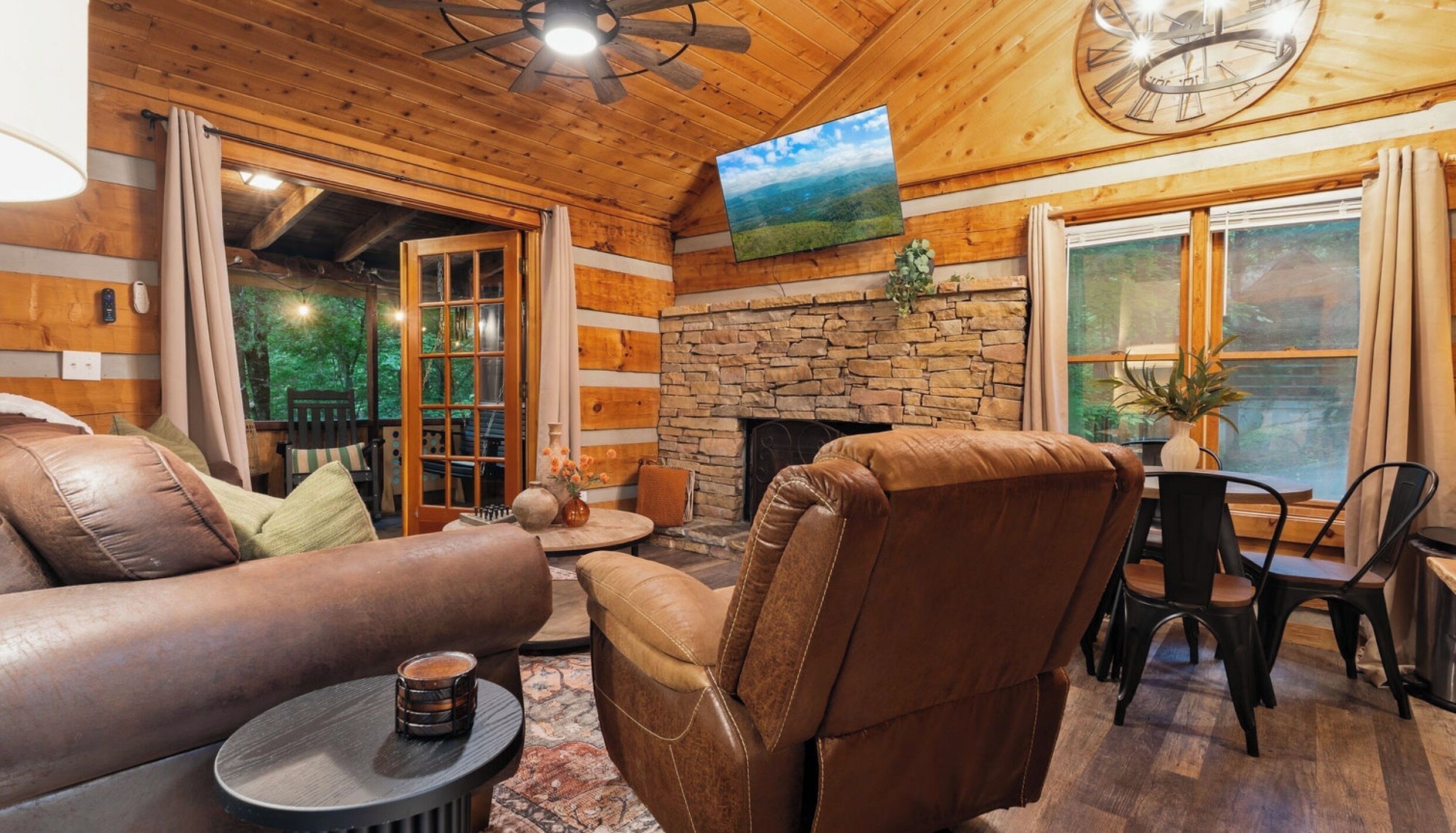 A cozy cabin interior with a stone fireplace, leather furniture, and a large window with a view of the outdoors.