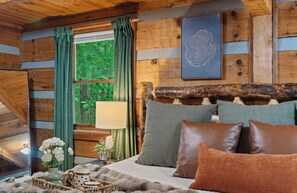 Un dormitorio de cabaña de madera con una cama, almohadas, una mesita de noche, una lámpara y una ventana con cortinas.