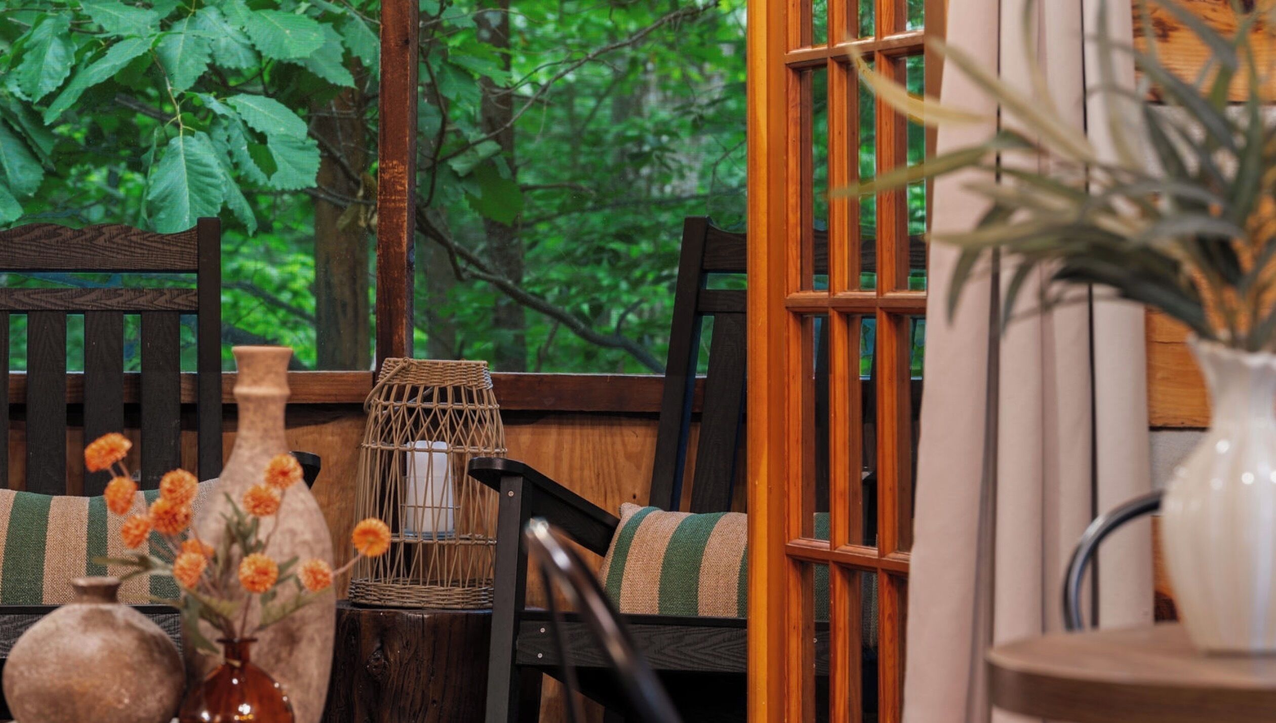 A wooden chair with a woven basket and a candle, a wooden table with a vase of flowers, and a view of greenery through a window.