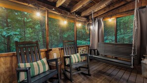 Une véranda en bois avec deux fauteuils à bascule et un banc, de grandes fenêtres donnant sur la forêt et des guirlandes lumineuses.