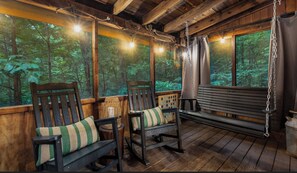 Un porche de madera con dos mecedoras y un banco, ventanas amplias con vistas al bosque y luces colgantes.