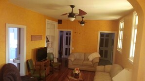 Un salon avec un canapé, un téléviseur et un ventilateur de plafond. Des portes donnent accès aux autres pièces.